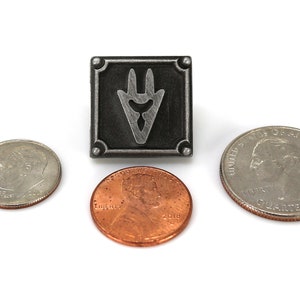 Op de afbeelding: Een zilverkleurige metalen pin met een zwarte achtergrond en een gestileerd zilverkleurig symbool. De pin is ongeveer zo groot als een Amerikaanse kwart.
