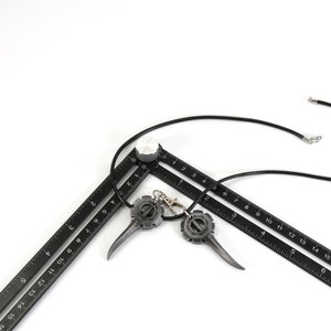 May include: A black leather necklace with two metal pendants shaped like a gear with a pointed tip. The necklace is laying on top of two black rulers.