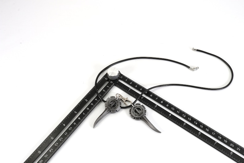 May include: A black leather necklace with two metal pendants shaped like a gear with a pointed tip. The necklace is laying on top of two black rulers.