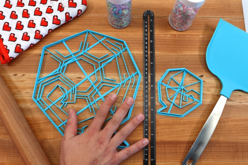 May include: Two blue plastic cookie cutters in the shape of snowflakes on a wooden surface. The larger cutter has a more intricate design than the smaller cutter.