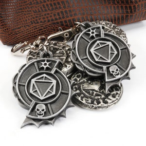 May include: A silver metal keychain with a dice symbol and a skull design. The keychain is attached to a brown leather pouch.