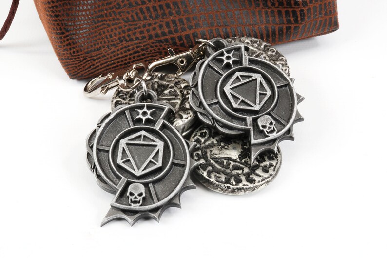 May include: A silver metal keychain with a dice symbol and a skull design. The keychain is attached to a brown leather pouch.