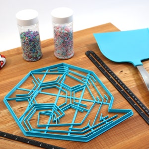 May include: A blue snowflake cookie cutter, two jars of colorful sprinkles, a black ruler, a blue spatula, and a red and white oven mitt on a wooden surface.