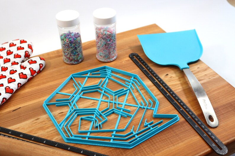 May include: A blue snowflake cookie cutter, two jars of colorful sprinkles, a black ruler, a blue spatula, and a red and white oven mitt on a wooden surface.