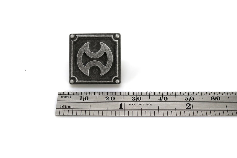 May include: A silver-coloured metal pin with a black and silver design. The design is a stylised symbol with two circles and a line in the middle. The pin is approximately 2 cm in diameter.