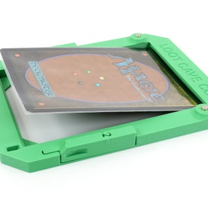 Op de afbeelding: Groene plastic kaarthouder met een Magic: The Gathering kaart erin. De houder heeft een hendel aan de zijkant en de tekst "LOOT CAVE CO." bovenaan.
