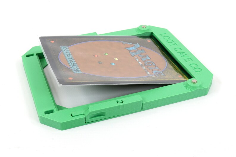 Op de afbeelding: Groene plastic kaarthouder met een Magic: The Gathering kaart erin. De houder heeft een hendel aan de zijkant en de tekst "LOOT CAVE CO." bovenaan.