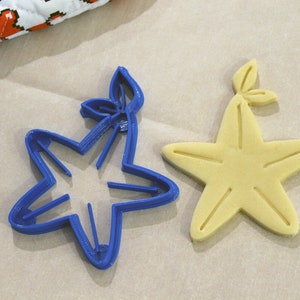 May include: A blue plastic cookie cutter shaped like a starfish with a leaf on top, next to a baked cookie in the shape of a starfish with a leaf on top.