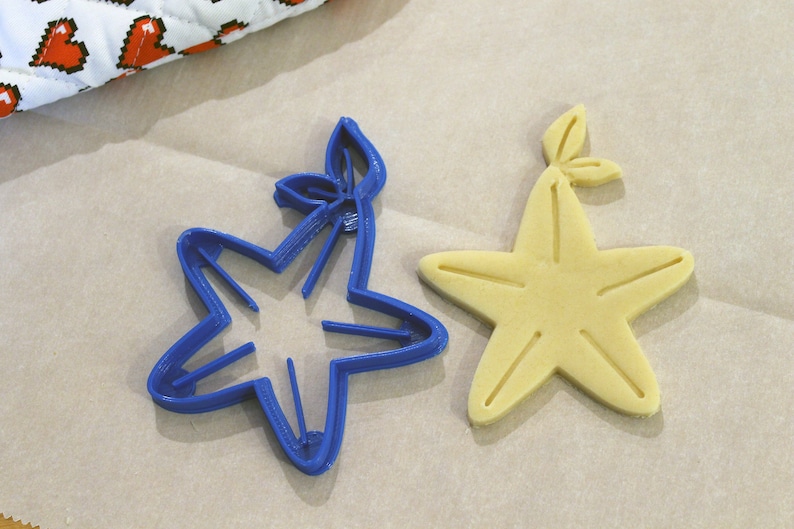 May include: A blue plastic cookie cutter shaped like a starfish with a leaf on top, next to a baked cookie in the shape of a starfish with a leaf on top.
