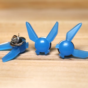 Peut inclure: Trois &eacute;pingles en plastique bleu en forme de symbole Triforce de la s&eacute;rie de jeux vid&eacute;o The Legend of Zelda. Les &eacute;pingles sont sur une surface en bois.