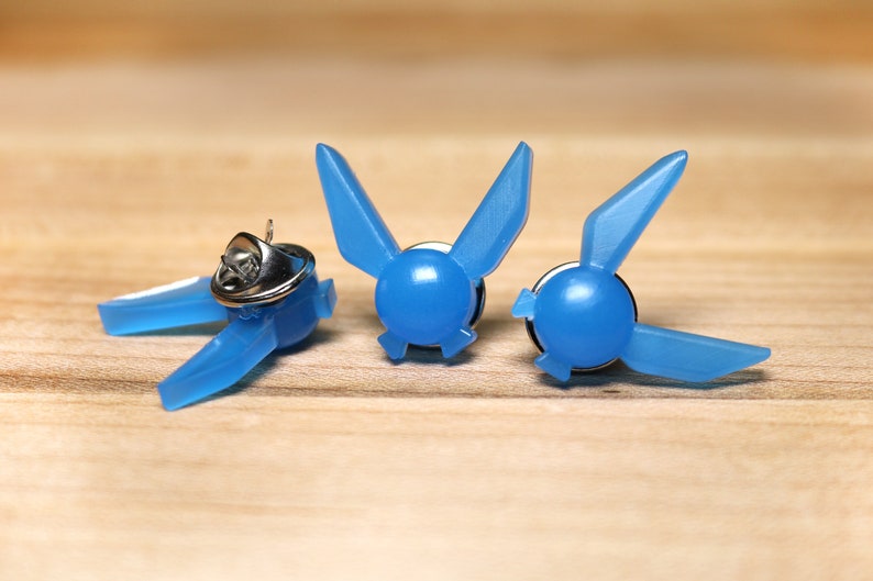 Peut inclure: Trois &eacute;pingles en plastique bleu en forme de symbole Triforce de la s&eacute;rie de jeux vid&eacute;o The Legend of Zelda. Les &eacute;pingles sont sur une surface en bois.