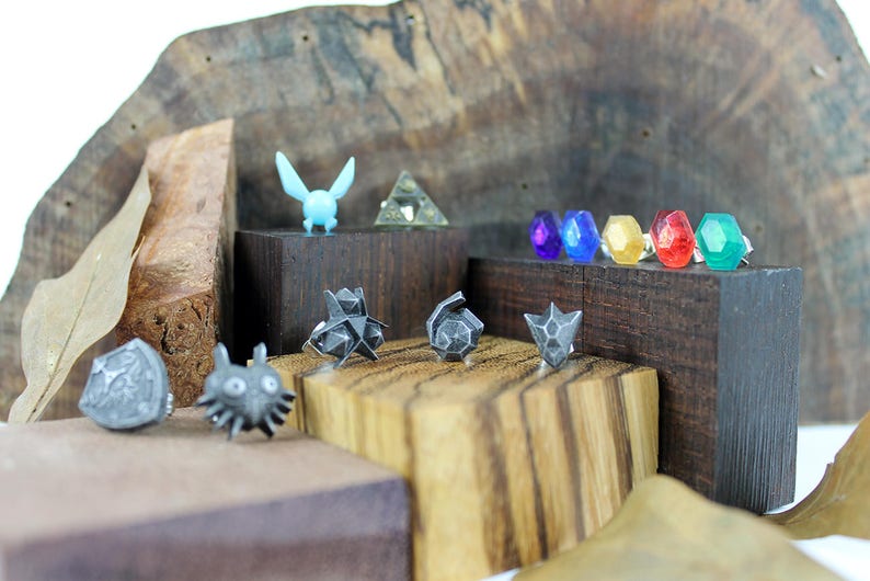 May include: A collection of small, silver-colored, 3D printed figurines, including a shield, a creature with large eyes, a triangular shape, and a winged creature. The figurines are displayed on small wooden blocks. There are also six small, colorful, gem-like objects on a wooden block.