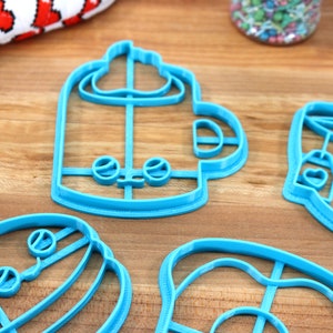 May include: Four blue plastic cookie cutters shaped like a coffee cup, a donut, a hamburger, and a mug with whipped cream. The cutters are on a wooden surface.