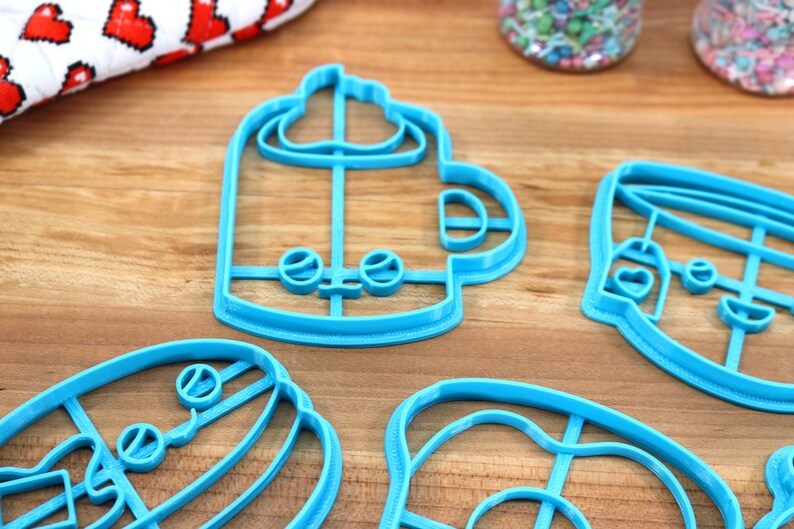 May include: Four blue plastic cookie cutters shaped like a coffee cup, a donut, a hamburger, and a mug with whipped cream. The cutters are on a wooden surface.