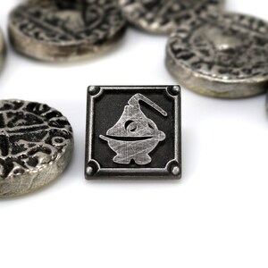 May include: A silver metal pin with a square design. The pin features a black background with a silver metal image of a stylized cauldron with a handle.