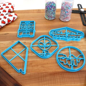 May include: A set of six blue plastic cookie cutters on a wooden surface. The cutters are shaped like a slot machine, a UFO, an alien, a kite, a cityscape, and a sign that says "Las Vegas Welcome".