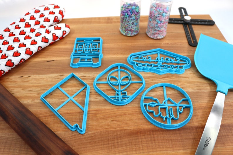 May include: A set of six blue plastic cookie cutters on a wooden surface. The cutters are shaped like a slot machine, a UFO, an alien, a kite, a cityscape, and a sign that says "Las Vegas Welcome".