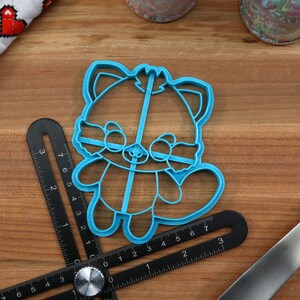 May include: A blue plastic cookie cutter shaped like a red panda. The cutter is on a wooden surface with a ruler and a spatula.