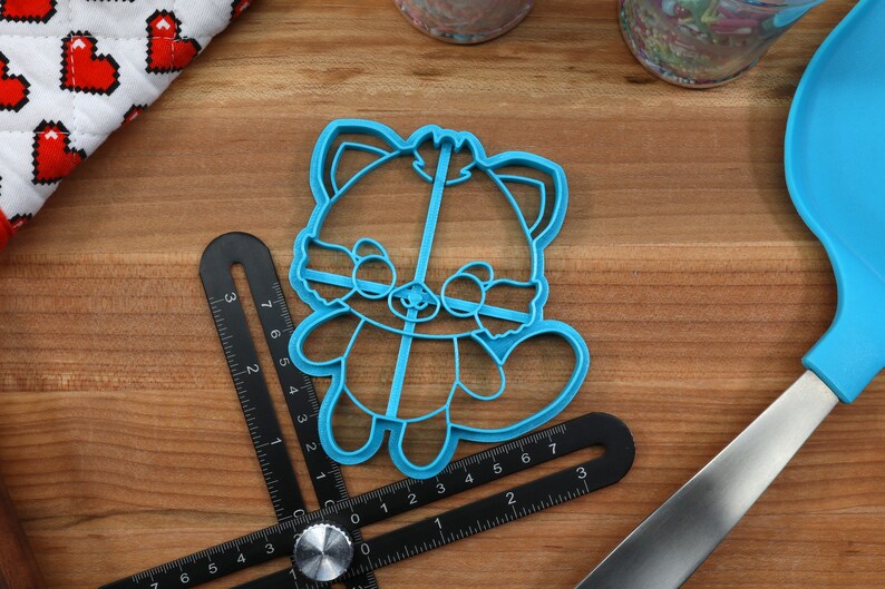 May include: A blue plastic cookie cutter shaped like a red panda. The cutter is on a wooden surface with a ruler and a spatula.