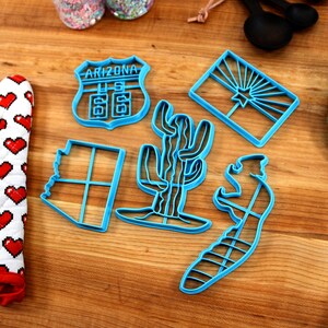 May include: A set of six blue plastic cookie cutters shaped like the state of Arizona, a cactus, a road sign that says "Arizona US 66", a saguaro cactus, a lizard, and the Arizona state flag.