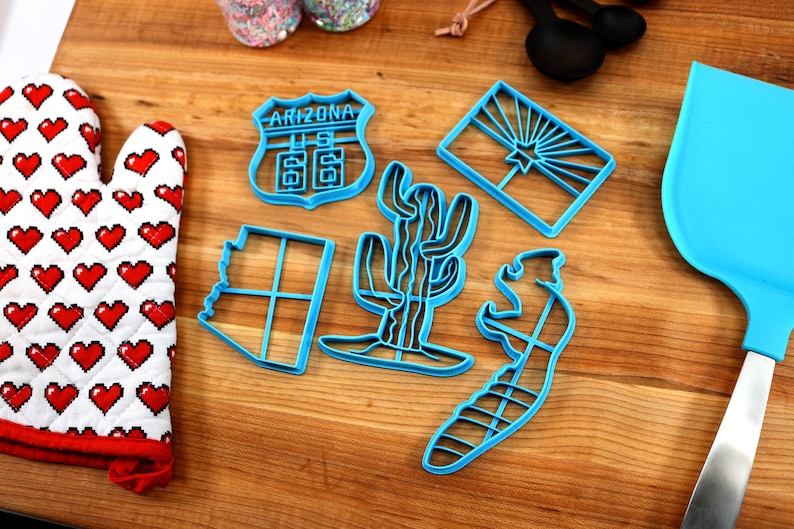 May include: A set of six blue plastic cookie cutters shaped like the state of Arizona, a cactus, a road sign that says "Arizona US 66", a saguaro cactus, a lizard, and the Arizona state flag.