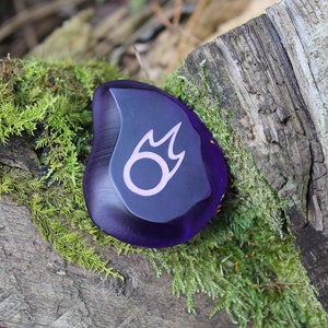 May include: A purple glass stone with a pink and black fire symbol etched on the surface.