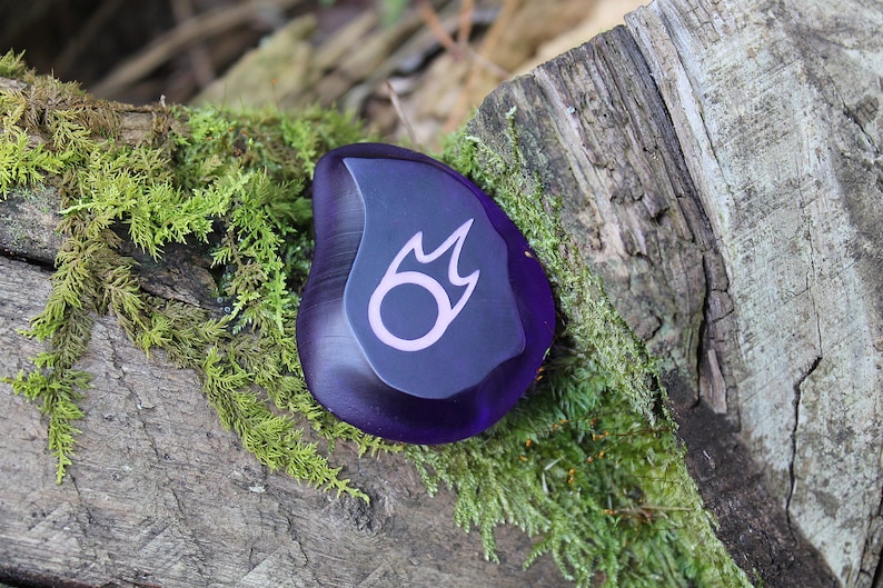 May include: A purple glass stone with a pink and black fire symbol etched on the surface.