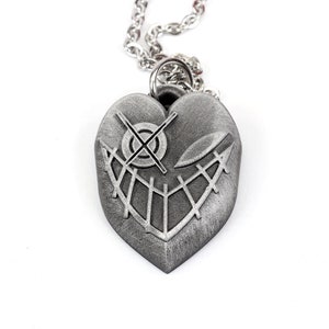May include: A silver heart-shaped pendant necklace with a detailed design of a stitched-up face with a cross for an eye.