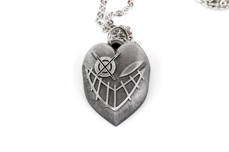 May include: A silver heart-shaped pendant necklace with a detailed design of a stitched-up face with a cross for an eye.