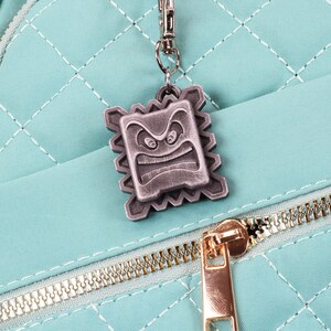 May include: A teal blue quilted backpack with a gold zipper and a silver metal charm featuring a cartoon face with an angry expression.