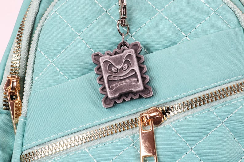 May include: A teal blue quilted backpack with a gold zipper and a silver metal charm featuring a cartoon face with an angry expression.
