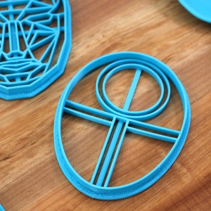 May include: A set of blue plastic cookie cutters, including a face, a circle with a cross inside, and an oval with a circle and cross inside.