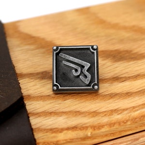 May include: A silver metal pin with a stylized design of a wing inside a square frame. The pin is on a wooden surface.