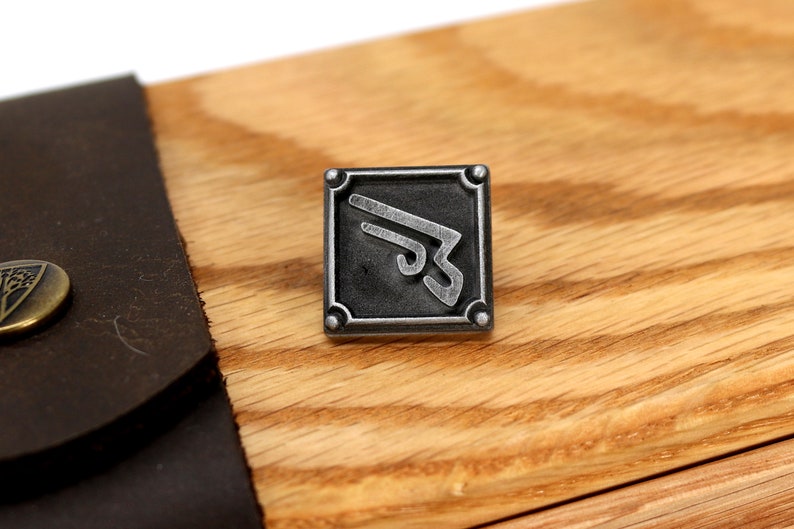 May include: A silver metal pin with a stylized design of a wing inside a square frame. The pin is on a wooden surface.