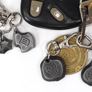 Może przedstawiać: Srebrny brelok do kluczy z czterema metalowymi zawieszkami. Zawieszki mają kształt tarcz i mają wygrawerowane na nich r&oacute;żne symbole. Brelok do kluczy jest przymocowany do złotego breloka z wygrawerowanym napisem "SERVICE".