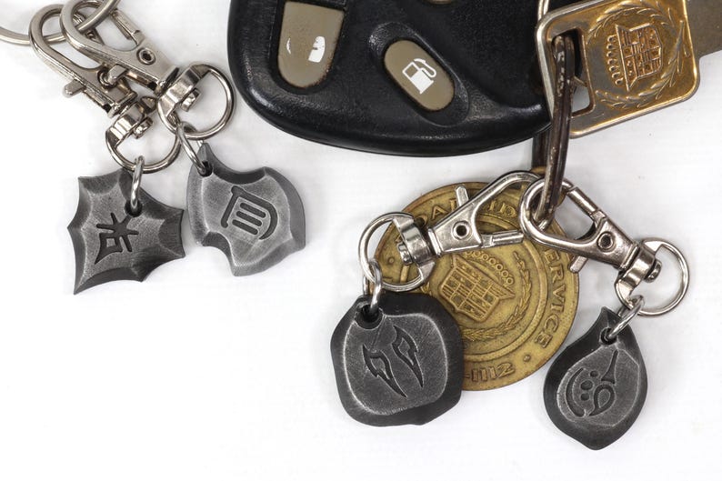 Może przedstawiać: Srebrny brelok do kluczy z czterema metalowymi zawieszkami. Zawieszki mają kształt tarcz i mają wygrawerowane na nich r&oacute;żne symbole. Brelok do kluczy jest przymocowany do złotego breloka z wygrawerowanym napisem "SERVICE".
