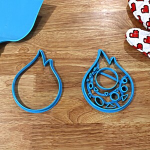 May include: Two blue plastic cookie cutters shaped like flames. One cutter is simple, while the other has a more intricate design with circles and triangles.