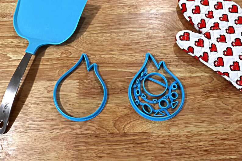 May include: Two blue plastic cookie cutters shaped like flames. One cutter is simple, while the other has a more intricate design with circles and triangles.