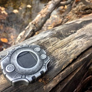 Puede incluir: Un colgante de metal plateado con un dise&ntilde;o circular y un cuadrado negro en el centro. El colgante est&aacute; sobre una pieza de madera desgastada.