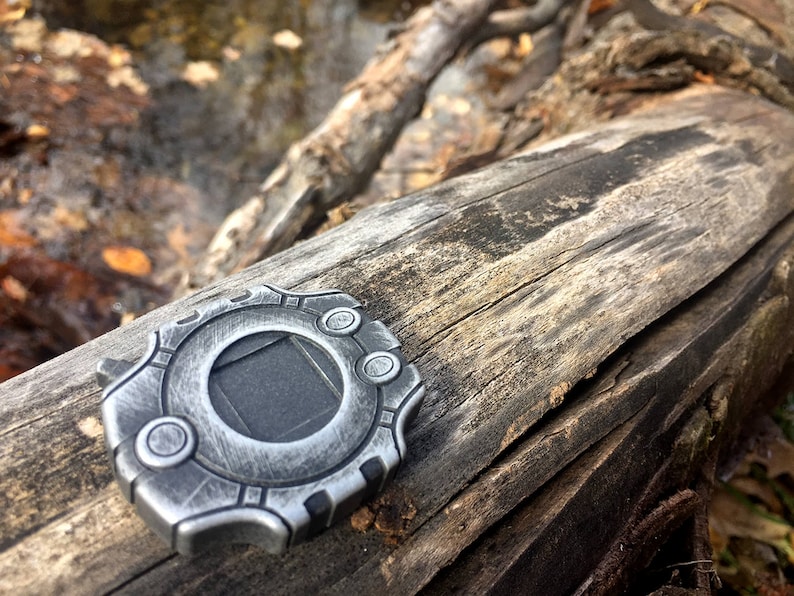 Puede incluir: Un colgante de metal plateado con un dise&ntilde;o circular y un cuadrado negro en el centro. El colgante est&aacute; sobre una pieza de madera desgastada.