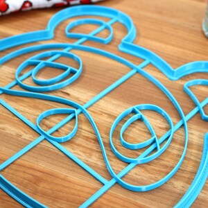 May include: A blue plastic cookie cutter shaped like a cartoon animal face. The cutter has a detailed design with multiple layers.