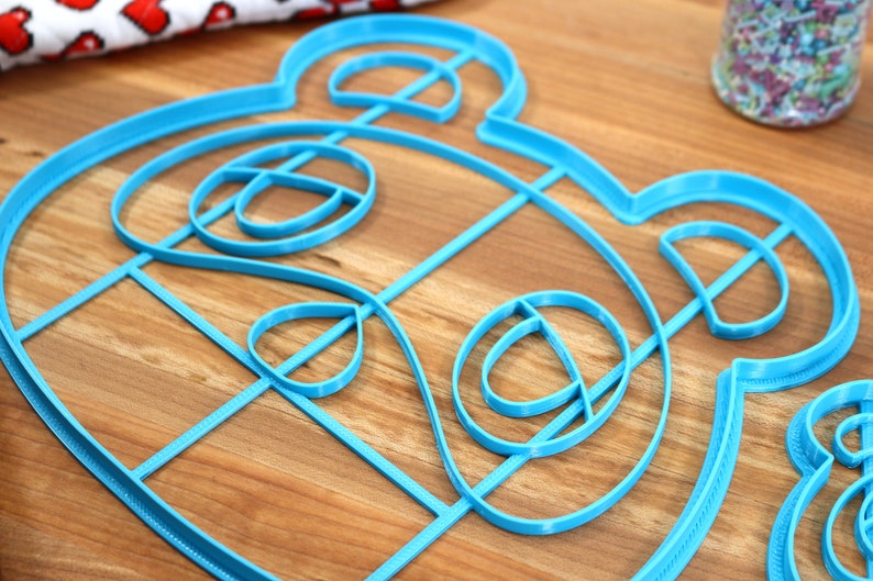 May include: A blue plastic cookie cutter shaped like a cartoon animal face. The cutter has a detailed design with multiple layers.