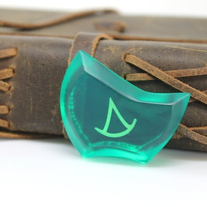 May include: A green, translucent,  dice-shaped object with a white symbol of a sailboat on it. The object is resting on a brown leather-bound journal.