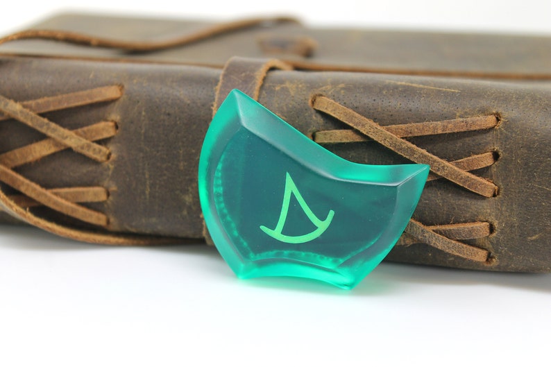 May include: A green, translucent,  dice-shaped object with a white symbol of a sailboat on it. The object is resting on a brown leather-bound journal.