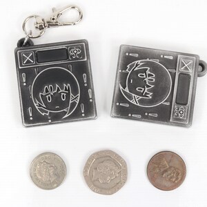 May include: Two silver keychains with a cartoon character on each. The keychains are shaped like a small square with a black screen and a silver button. The character on the keychain is a cartoon girl with black hair and a white face. The keychains are on a white background with three silver coins.