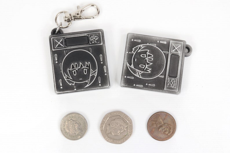 May include: Two silver keychains with a cartoon character on each. The keychains are shaped like a small square with a black screen and a silver button. The character on the keychain is a cartoon girl with black hair and a white face. The keychains are on a white background with three silver coins.