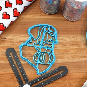 May include: A blue plastic cookie cutter in the shape of a person holding a barrel, with a ruler, spatula, and oven mitt with red hearts. Sprinkles and a rolling pin are also visible.