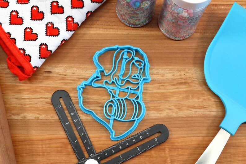 May include: A blue plastic cookie cutter in the shape of a person holding a barrel, with a ruler, spatula, and oven mitt with red hearts. Sprinkles and a rolling pin are also visible.