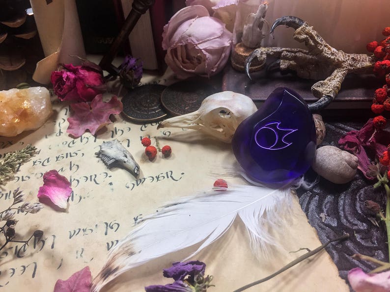 Op de afbeelding: Een donkerblauwe glazen hanger met een zilveren vlamontwerp, omringd door verschillende objecten, waaronder een vogelschedel, een veer en gedroogde bloemen.