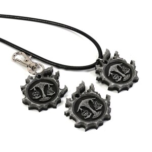 May include: A black leather necklace with a silver pendant featuring a stylized design. The pendant is a circular gear with a tree and scales in the center. The necklace comes with two additional pendants, one attached to a silver key chain.